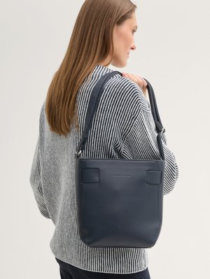 Femme aux longs cheveux bruns portant un pull rayé noir et blanc et un sac à bandoulière en cuir bleu marine.
