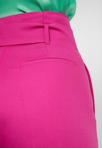 Ljust rosa byxor med en slät tygstruktur, med en hög midja och diskreta bakfickor, matchat med en grön topp.