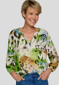 Frau lächelt, trägt eine bunte Bluse mit Tierdruck, Stern und "LOVE"-Schriftzug, kombiniert mit blauen Jeans, Hände in den Taschen, neutraler Hintergrund.