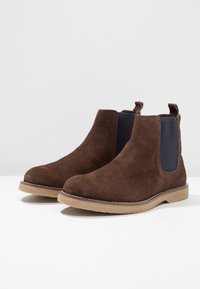 Braune Wildleder Chelsea-Stiefel mit schwarzen elastischen Seiteneinsätzen, einer abgerundeten Zehenform und einer tanfarbenen Gummi-Außensohle. Stichdetails entlang der Sohle.