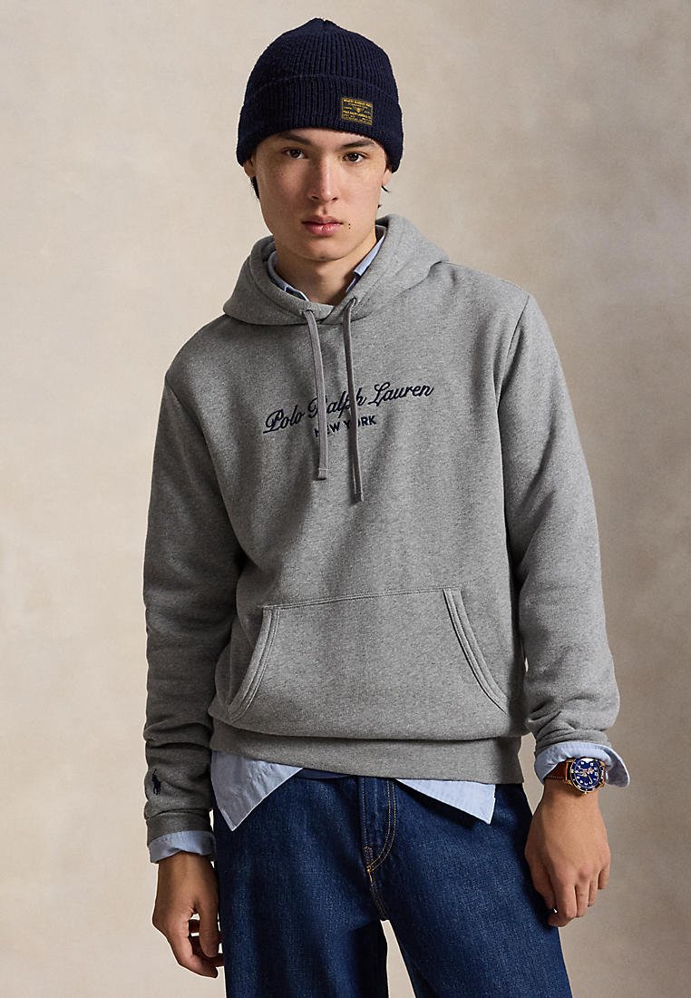 Modèle portant un bonnet en tricot bleu marine, un sweat à capuche gris Polo Ralph Lauren, un jean bleu et une montre-bracelet bleue, sur un fond de studio beige.