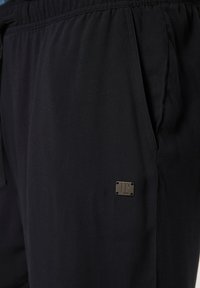 Pantaloni sportivi neri con vita elasticizzata, tasche laterali e una piccola etichetta metallica sul lato sinistro. Tessuto dalla consistenza liscia.