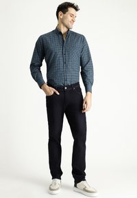 Camicia a maniche lunghe a quadri blu e verdi con bottone e taschino, abbinata a jeans scuri e sneakers chiare.