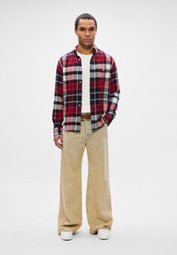 Hombre vestido con una camiseta blanca y una camisa de cuadros roja, negra y blanca, pantalones anchos beige, cinturón marrón y zapatillas blancas, de pie contra un fondo liso.