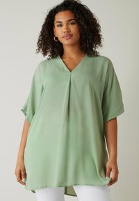 Lösa, kortärmade toppar i lätt grön tyg. Har en V-ringning och en veckdetalj fram. Slät textur, höftlång design.