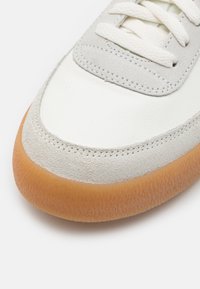 Zbliżenie białego i jasnoszarego czubka trampka z teksturowaną brązową gumową podeszwą i białymi sznurówkami na białym tle.