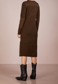 Abito lavorato a maglia marrone con maniche lunghe, silhouette aderente e dettagli delle cuciture. Presenta un accento con un anello sul retro. Abbinato a stivali cowboy neri.