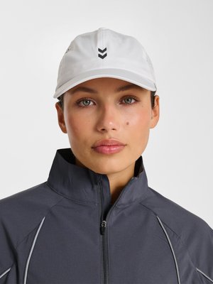 Kvinde med lys hud, grønne øjne og et skønhedsplet, iført en hvid kasket og en mørk lynlåstrøje, der vender fremad.