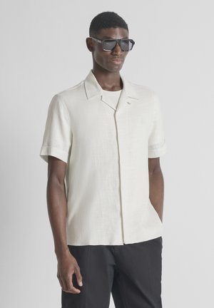 Homme portant des lunettes de soleil carrées noires, une chemise blanche à manches courtes avec col et un pantalon noir, debout devant un fond gris clair uni.