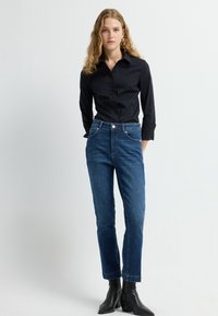 Camisa negra de botones, jeans de mezclilla y botines negros. Los jeans presentan un diseño clásico de cinco bolsillos, un corte ligeramente afinado y un color azul medio.