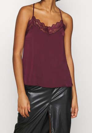 Frau trägt ein ärmelloses bordeauxfarbenes Oberteil mit Spitzenbesatz und dünnen Trägern, kombiniert mit einem schwarzen Lederrock mit Frontschlitz.