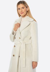 Abrigo blanco de doble pecho con una tela suave y texturizada, grandes solapas y un cinturón atado. Presenta botones marrones en la parte delantera.