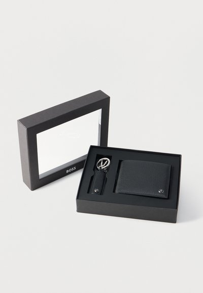Portefeuille en cuir noir texturé et porte-clés assorti avec anneau métallique dans une boîte cadeau noire ouverte avec couvercle transparent.