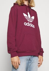 Bordeaux hoodie med vit Adidas-logga, dragsko på huvan och framficka. Tillverkad av mjukt material med ribbade ärmslut och nederkant.