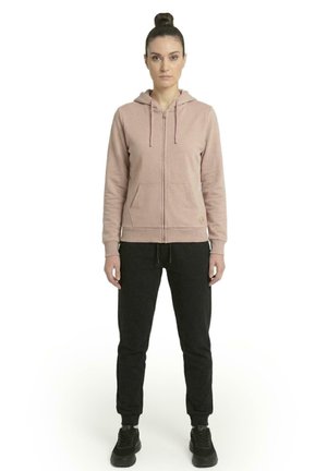 Une femme porte un sweat à capuche rose pâle zippé avec des cordons de serrage et un pantalon de jogging noir, sur un fond blanc uni.