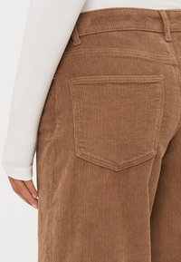 Bruine corduroy broek met een brede pijp, voorzien van een achterzak en een verticale ribbelstructuur. De tailleband is gestikt voor extra detail.