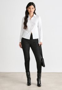 Bílá košile s límcem, spárovaná s černými skinny džíny. Modelka drží malou černou kabelku přes rameno a nosí černé kotníkové boty.