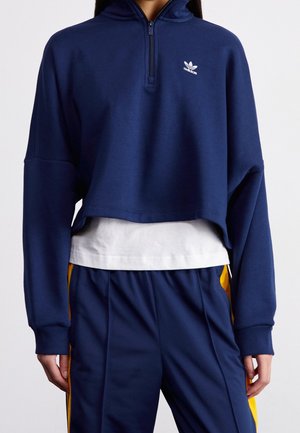 Persoon die een marineblauw cropped Adidas half-zip sweatshirt draagt over een wit shirt met marineblauwe en gele trainingsbroek.
