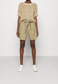 Vaaleanbeige neulotun villapaidan, jossa on lyhyet hihat, yhdistettynä vaaleankhakiin shortseihin, joissa on solmittava vyötärö. Valkoiset tennislenkkarit täydentävät asun.