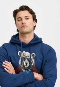 Felpa con cappuccio blu navy realizzata in tessuto morbido, caratterizzata da una grafica di un orso con occhiali da sole. Il design include cordini e una tasca frontale a marsupio.