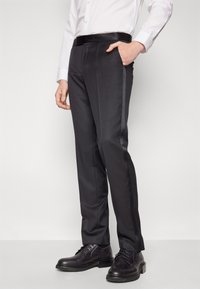 Calças pretas de corte impecável, com um tecido suave, bolsos laterais e um acabamento em seda nas costuras, combinadas com sapatos pretos.