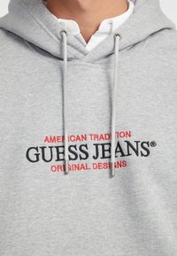 Felpa grigia realizzata in morbido tessuto, con ricamo frontale in rosso e nero: "AMERICAN TRADITION GUESS JEANS ORIGINAL DESIGNS."