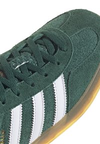 Vihreät samettiset lenkkarit valkoisilla raidallisilla yksityiskohdilla, tasaisilla nauhoilla ja kumisella pohjalla, joissa on teksturoitu päällinen ja ommeltuja yksityiskohtia.