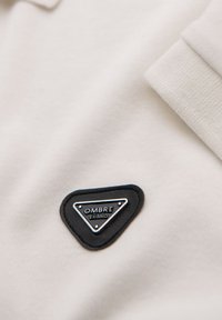 Pikétröja i ljusbeige tyg med en svart triangulär patch med silverdetaljer som läser "OMBRE EXÍMIO." Texturerad krage synlig.