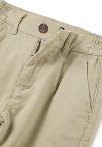 Beige corduroy broek met een rits- en knoopsluiting, elastische tailleband en zijzakken. De textuur is ribbelig met een zachte afwerking.