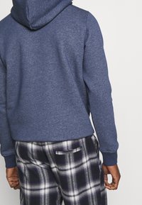 Sudadera con capucha de color azul marino con puños acanalados, combinada con pantalones de franela a cuadros en blanco y negro que cuentan con un bolsillo trasero. Tejido suave y texturizado.