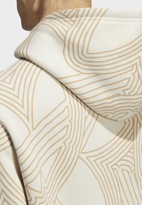 Mikina s kapucí vyrobená z krémové látky, která má zakřivené béžové vzory. Měkká textura s viditelným švem na kapuci.