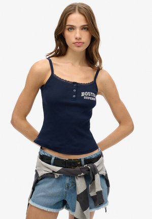 Marineblauwe ribbel tanktop met kantafwerking, voorzien van knopen en de tekst "BOSTON SUPERDRY", gecombineerd met denim shorts en een grijze sweatshirt die om de taille is gebonden.