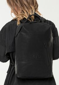 Frau mit schulterlangem Haar, trägt eine schwarze Jacke und einen glatten schwarzen Rucksack mit vorderer Reißverschlusstasche und geprägt präsentiertem Logo.