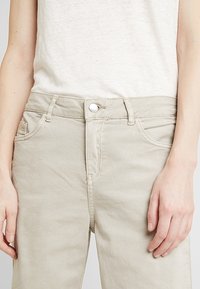 Beiget puuvillahousut suoralla lahkeella, joissa on taskut ja edessä nappisuljenta. Kangas on pehmeän tuntuinen ja hienovaraisesti kiiltävä.