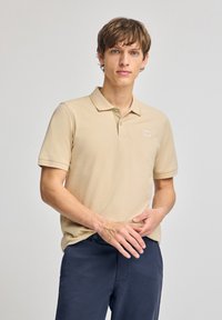 Beige kortärmad pikétröja med krage, med en liten broderad logotyp på vänster bröst. Bärs tillsammans med marinblå byxor.