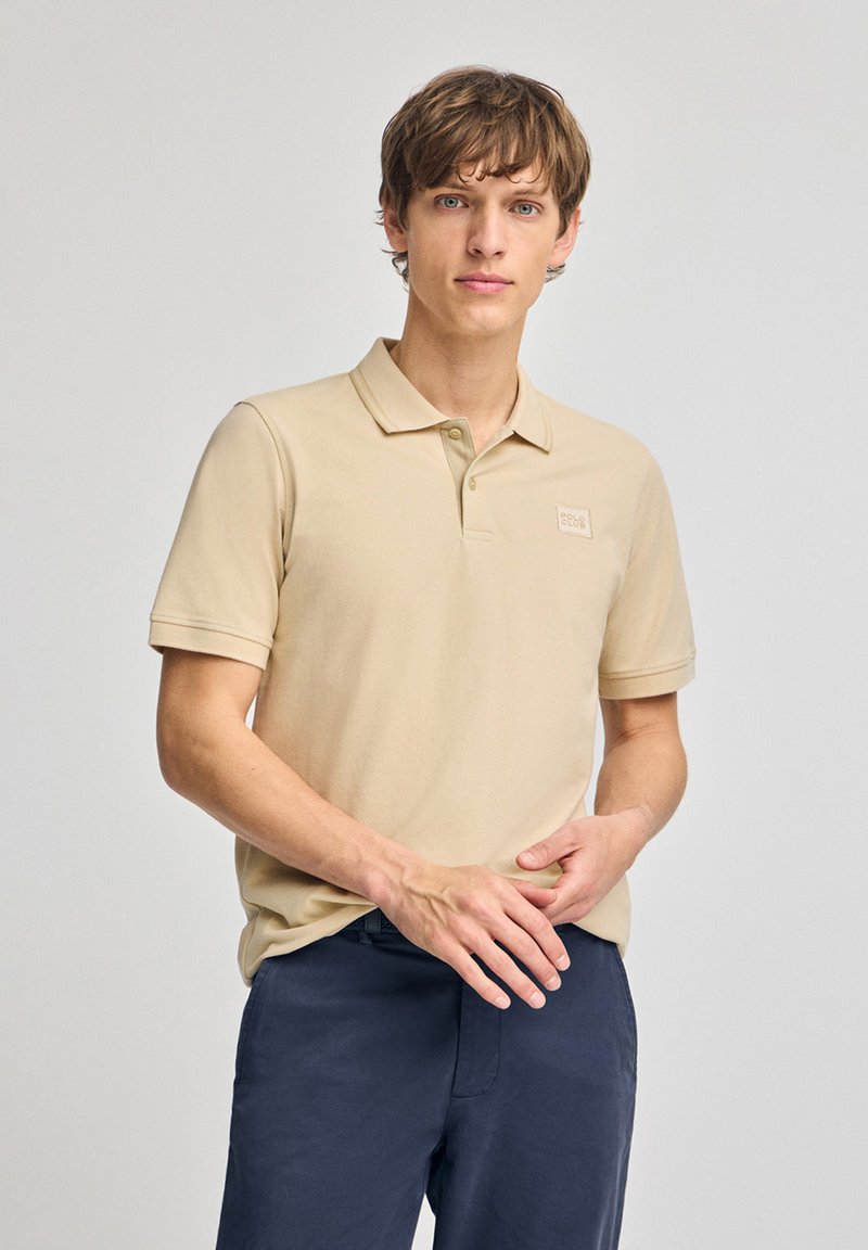 Beige kortärmad pikétröja med krage, med en liten broderad logotyp på vänster bröst. Bärs tillsammans med marinblå byxor.