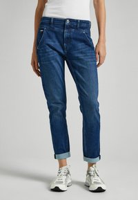 Vaqueros de denim en azul oscuro con corte recto, con bolsillos frontales, costuras claras y puños doblados. Llevados con zapatillas blancas.