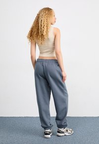 Joven mujer con cabello largo y rizado de espaldas, lleva una camiseta sin mangas beige, pantalones de chándal grises y zapatillas blancas sobre una alfombra azul.