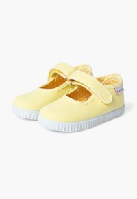 Pantofi din pânză galbenă cu talpă din cauciuc alb, dotat cu o curea Velcro și căptușeală interioară în carouri. Vârf rotund și detalii de design minime.