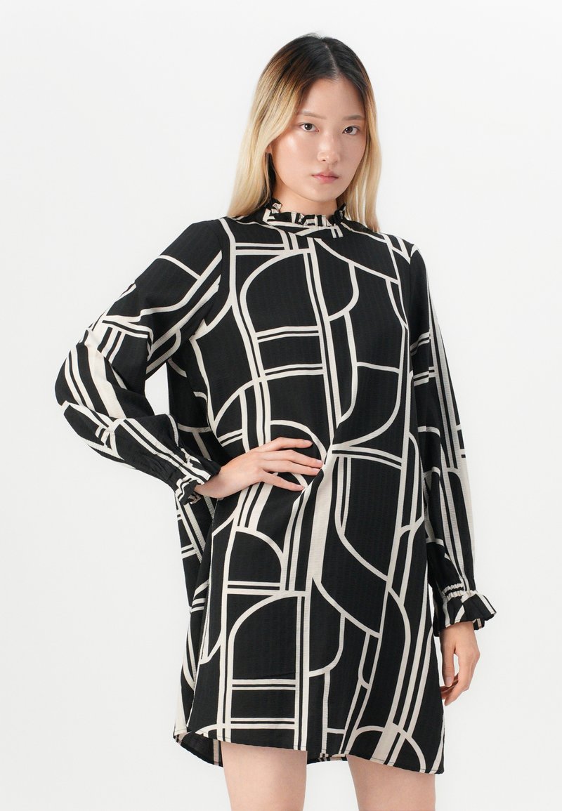 Mujer de pie con la mano en la cadera, vistiendo un vestido negro con líneas geométricas blancas y largas mangas arrugadas, contra un fondo blanco.