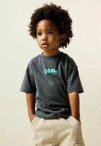 Szürke, rövid ujjú póló, melyen a mellrészen 3D türkiz "COOL" felirat díszeleg, világos bézs rövidnadrággal párosítva. Simogató pamut anyagból.