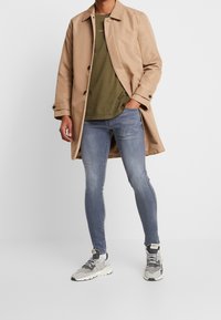 Abrigo trench beige sobre una camiseta verde oliva, combinado con jeans skinny de un azul claro y zapatillas grises con acentos negros y suelas blancas.