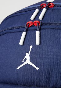 Navy backpack with textured fabric, featuring red zip pulls and white Jordan logo. Durable design includes multiple zippered compartments.