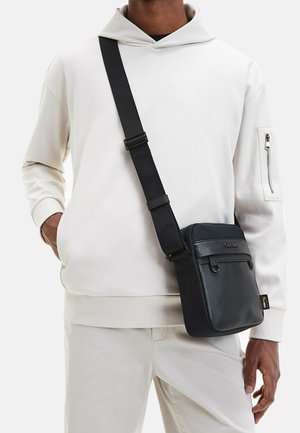 Persoon draagt een gebroken witte hoodie en broek, met een zwarte crossbodytas met verstelbare riem over de schouder.