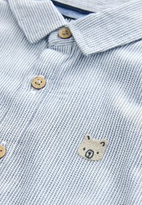 Chemise à rayures bleu clair avec des boutons en bois et un petit visage d'ours beige brodé sur la poitrine près des boutons.