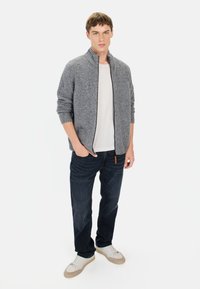 Grijze zip-up trui met een gestructureerd breiwerk, een wit T-shirt eronder, donkere jeans en witte sneakers. Opvallende ritsdetails en een bruine accent.