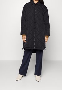 Osoba oblečená v černém prošívaném dlouhém kabátu se zipem vpředu, námořnických širokých kalhotách a bílých pletených kotníkových botách, stojící na bílém pozadí.