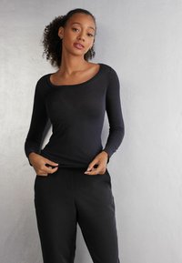 Schwarzes Langarmoberteil mit einem weiten Ausschnitt, aus einem glatten Stoff gefertigt, kombiniert mit schwarzen Hosen. Das Modell passt den Saum an, während es posiert.