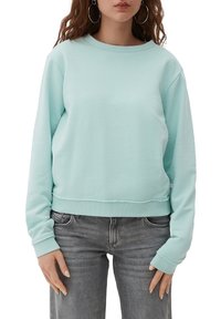 Mujer vestida con sudadera de manga larga azul claro y jeans gris desgastado, de pie con las manos relajadas a los lados.