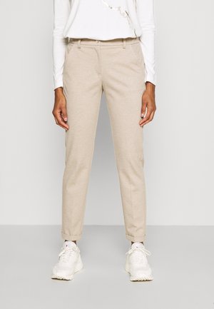 Person trägt beige gerade geschnittene Hosen mit umgeschlagenen Säumen und weißen Sneakers, steht vor einem schlichten hellen Hintergrund.